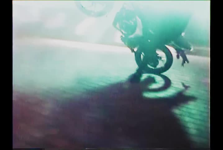 Gritty, vintage-style footage captures a motorcyclist doing a wheelie on a brick street amidst thick smoke at night
