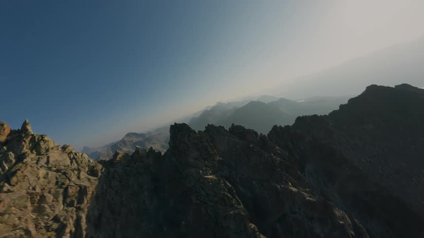 An FPV drone soars backwards along a dramatic, craggy mountain ridge, emerging from haze into the bright morning sun over a vast alpine valley