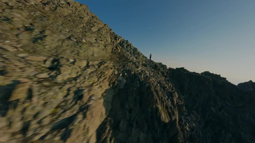 FPV drone footage follows a lone adventurer trekking along a steep and rocky mountain trail at high altitude