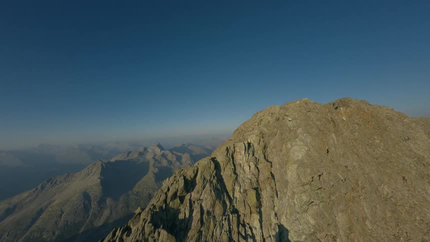 An aerial view flying along a rugged and rocky mountain ridge towards the summit against a clear blue sky