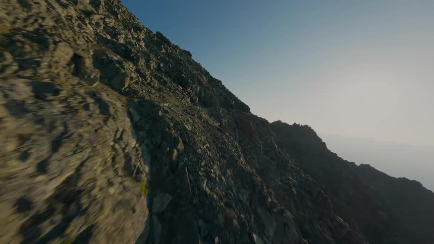 Thrilling FPV drone footage follows hikers navigating a narrow and rocky mountain ridge against a backdrop of a vast mountain range at sunrise