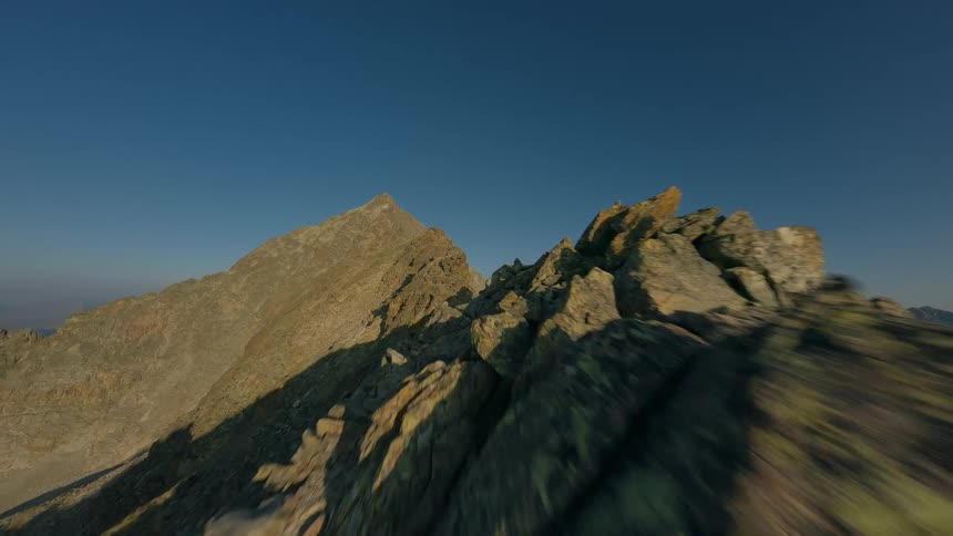 An exhilarating FPV drone flight soars along a narrow, rocky mountain ridge under a clear blue sky at sunrise