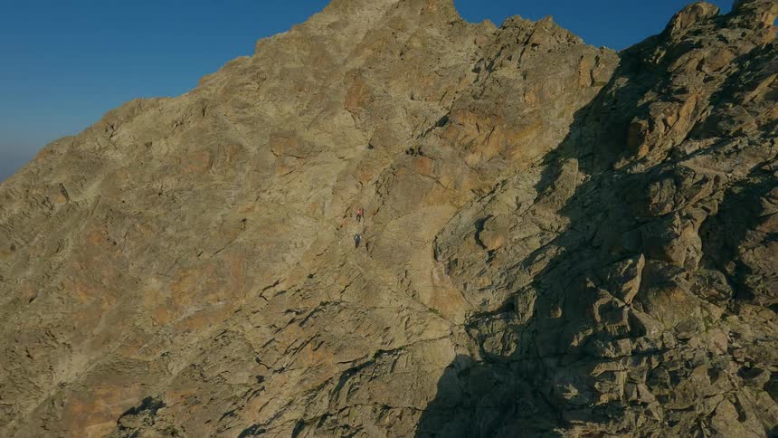 An FPV drone soars alongside a steep, rocky mountain ridge, revealing two climbers ascending the sunlit face of the peak