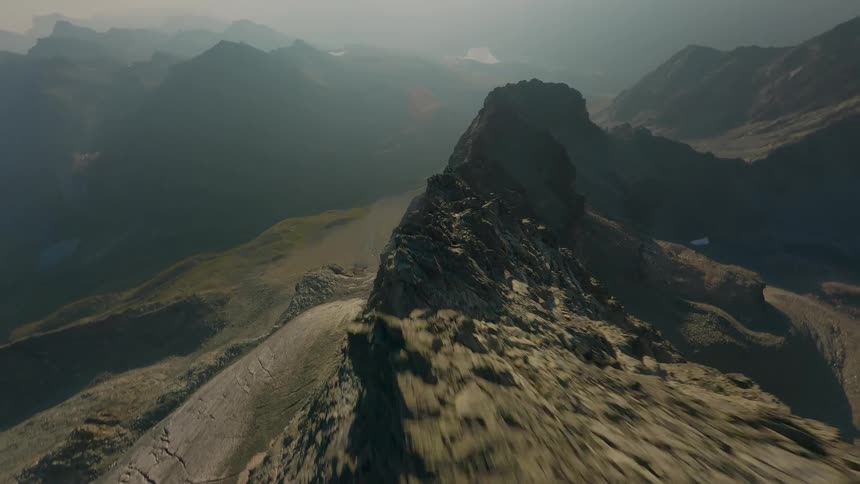 A thrilling FPV drone flight dives down the steep, rocky spine of a mountain range on a hazy day