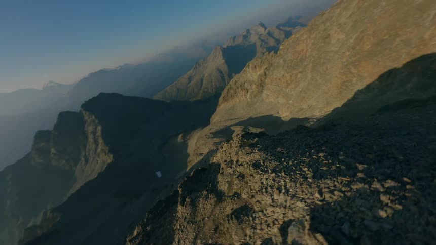 An exhilarating FPV drone flight follows a hiker traversing a steep and rocky alpine ridge in a vast mountain landscape
