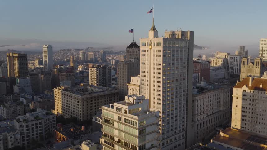A majestic aerial view captures the historic architecture of San Francisco's skyline, bathed in the warm light of sunrise with fog lingering over the distant hills