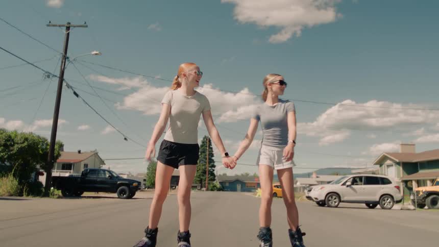 Two happy teenage girls rollerblade hand-in-hand towards the camera in slow motion on a sunny suburban street