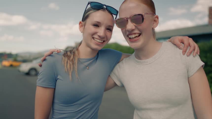 Two happy young women laugh and smile for the camera while embracing on a sunny suburban street