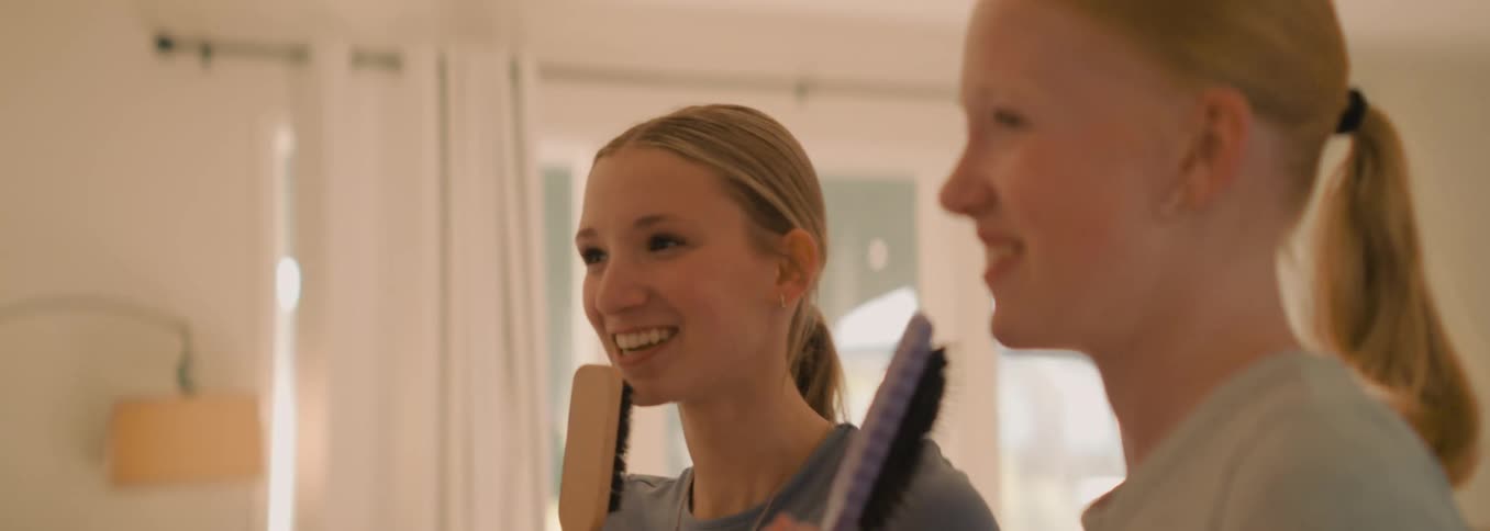 Two joyful teenage girls laugh and sing together while using hairbrushes as microphones in their room