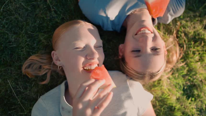 A delightful top-down view of two happy teenage girls laughing and eating juicy watermelon while lying on the green grass on a sunny day