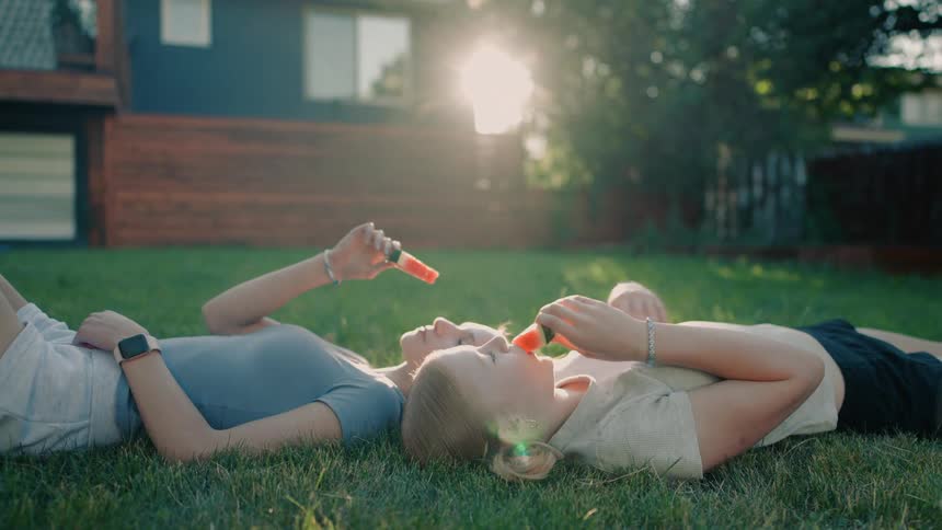 Slow motion shot of two teenage girls laying on the grass in a sunny backyard eating watermelon