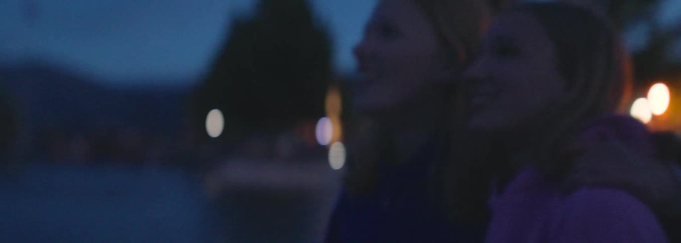 Two smiling teenage girls hug each other as they watch a beautiful fireworks display at night