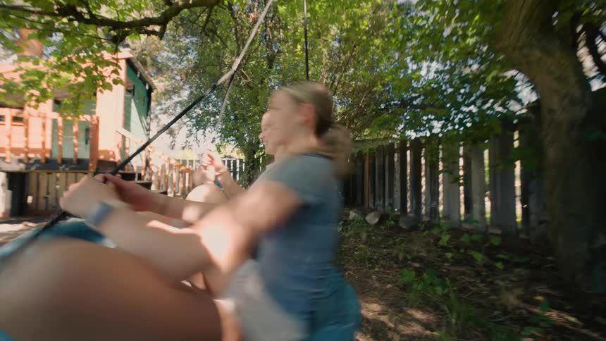 Two happy teenage girls spin and laugh together on a round swing in a sunny backyard
