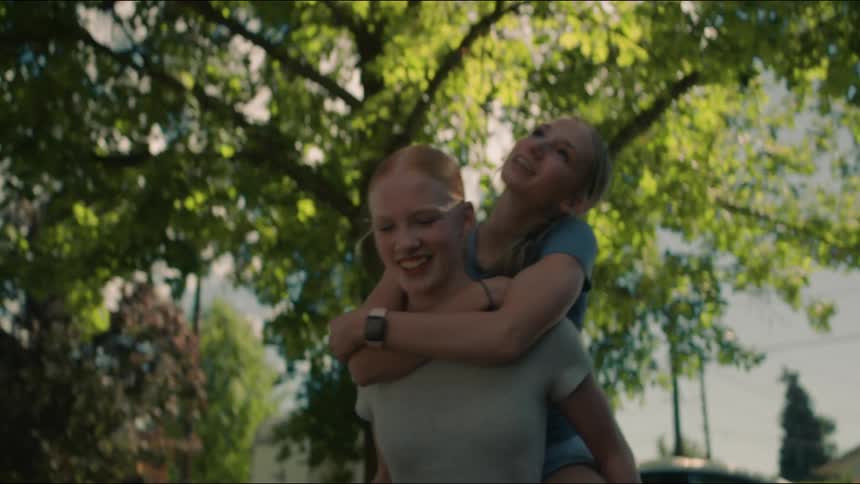 A smiling girl gives her friend a piggyback ride as they walk through their neighborhood on a sunny summer day
