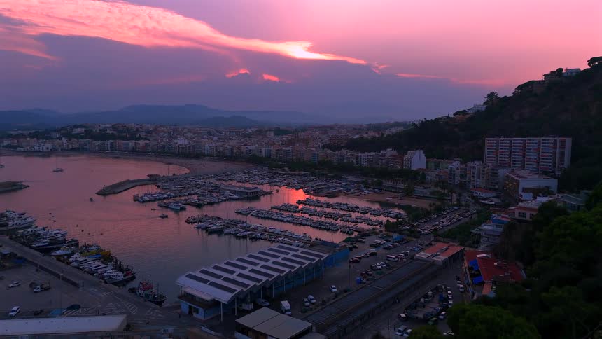 An enchanting aerial view captures a coastal town and marina filled with boats, bathed in the stunning pink and purple hues of sunset