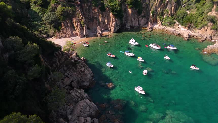 An idyllic aerial view shows numerous boats and yachts anchored in the clear turquoise water of a secluded cove surrounded by rocky cliffs