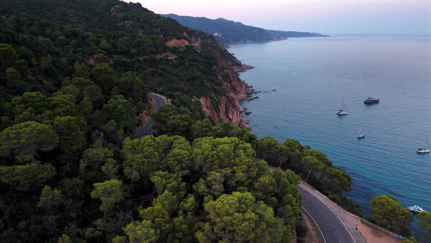 Drone flies backwards over a winding coastal road along a forested cliffside, revealing the tranquil sea with anchored boats at sunset