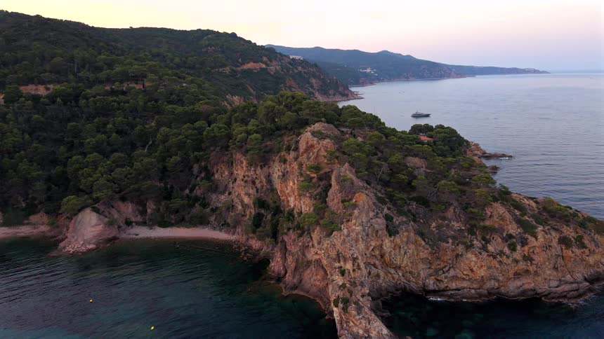 A slow aerial pullback reveals a stunning, tree-covered rocky coastline with a hidden cove and calm turquoise sea at sunset