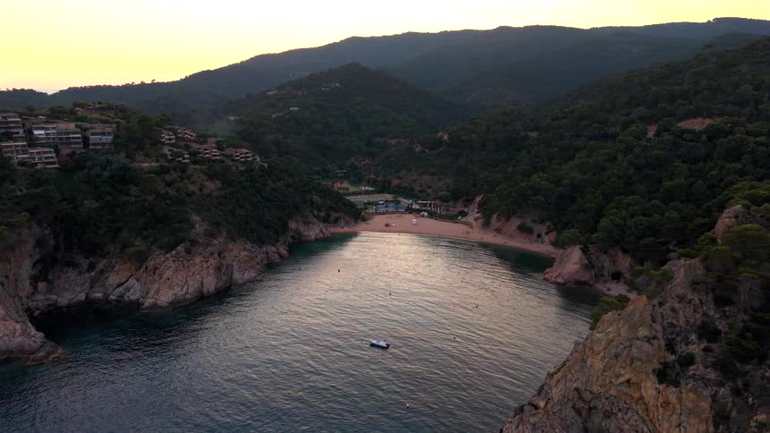 A tranquil aerial pullback shot reveals a beautiful secluded beach cove surrounded by rocky cliffs and forested hills at sunset