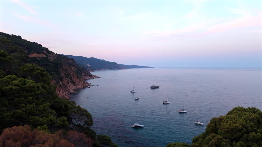 An aerial drone shot flies over a lush green forest to reveal a beautiful coastal bay with several boats anchored in the calm water at dusk