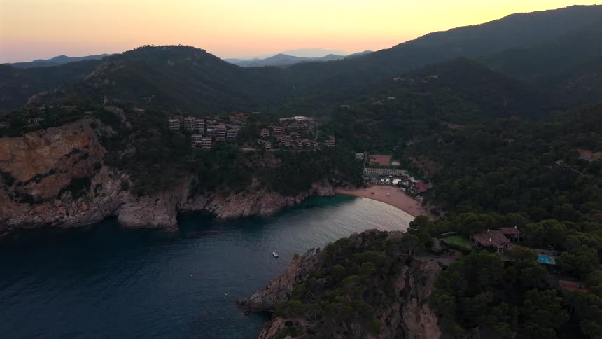A stunning aerial view of a secluded beach cove surrounded by lush green hills and rocky cliffs during a peaceful sunset