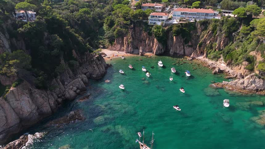 Aerial drone view slowly pulls back from a stunning secluded bay with crystal clear turquoise water, anchored yachts, and a hidden beach