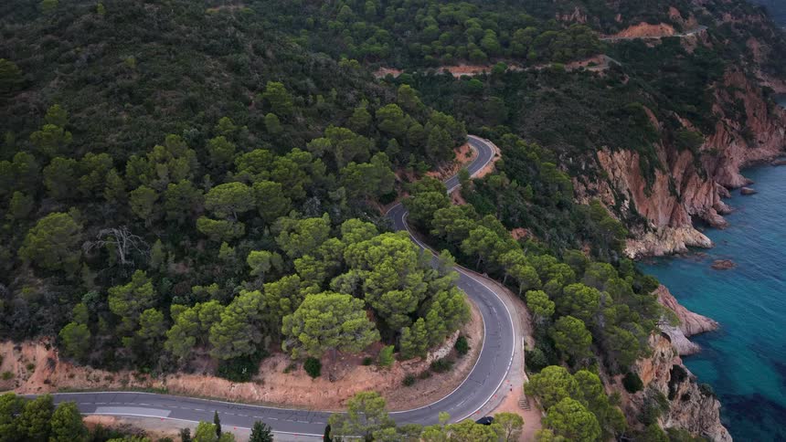 An aerial view shows cars navigating a winding coastal road carved into a steep, forested cliffside overlooking the turquoise ocean