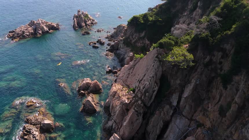 An aerial view reveals a stunning rocky coastline with clear turquoise water as a kayaker paddles towards a nearby marina on a sunny day