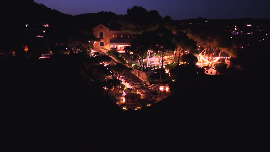 Aerial shot pulls back from a beautifully lit hillside villa event venue at night, revealing the surrounding landscape