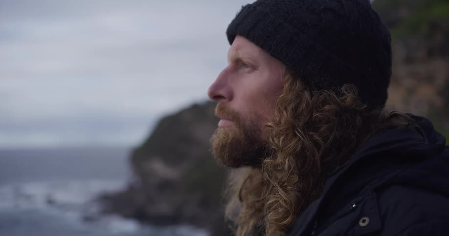 A pensive man with a long beard and curly hair contemplates the view from a rocky coastline on an overcast day