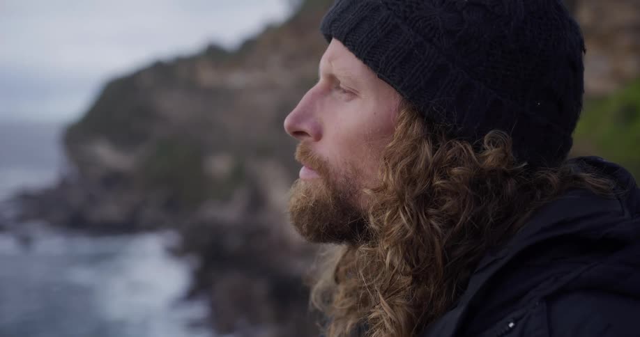 A slow-motion rack focus shot reveals a contemplative man with long hair and a beard looking out at the moody, rocky coastline
