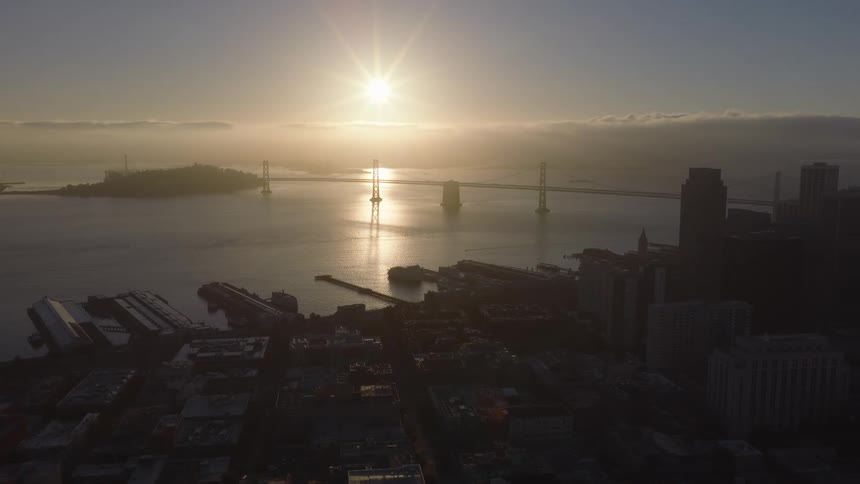 A serene aerial view shows the sun rising over the San Francisco Bay, silhouetting the city skyline and the iconic Bay Bridge in a hazy golden light