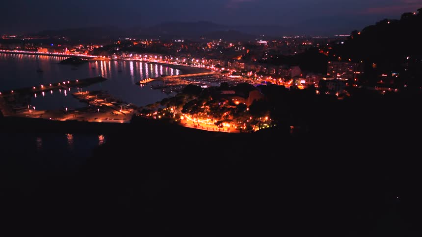 A mesmerizing aerial view captures a brightly lit coastal town at night, slowly revealing a festive event on a seaside hill next to a calm harbor