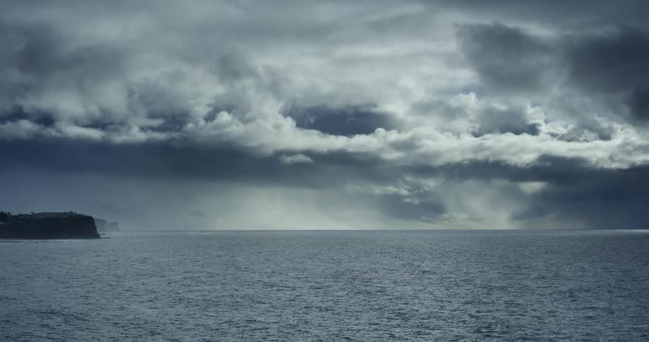 Sunlight breaks through dark, dramatic storm clouds to illuminate a rain shower falling over the ocean