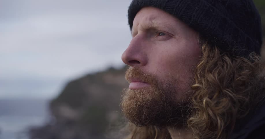 Close up of a pensive man with a beard and beanie looking out over a moody coastline