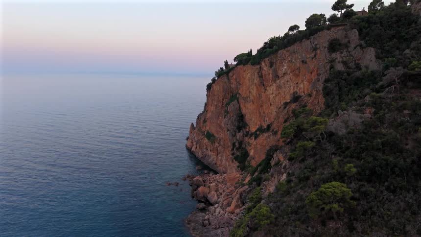 An aerial shot reveals a beautiful rocky cliff face covered in trees overlooking the calm sea at dusk