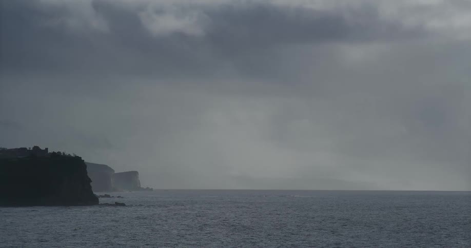 A calm gray ocean meets a foggy coastline under a heavy, overcast sky
