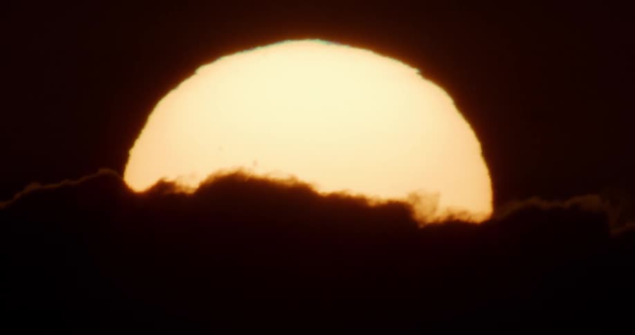 An extreme close-up shot of the bright yellow sun setting behind dark, silhouetted clouds