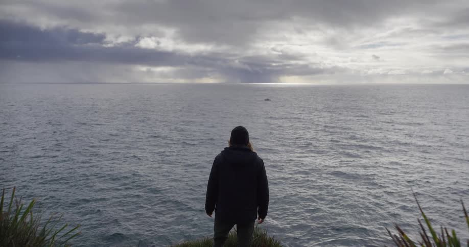 A lone figure stands on a cliff's edge, gazing out at the vast ocean under a dramatic, stormy sky