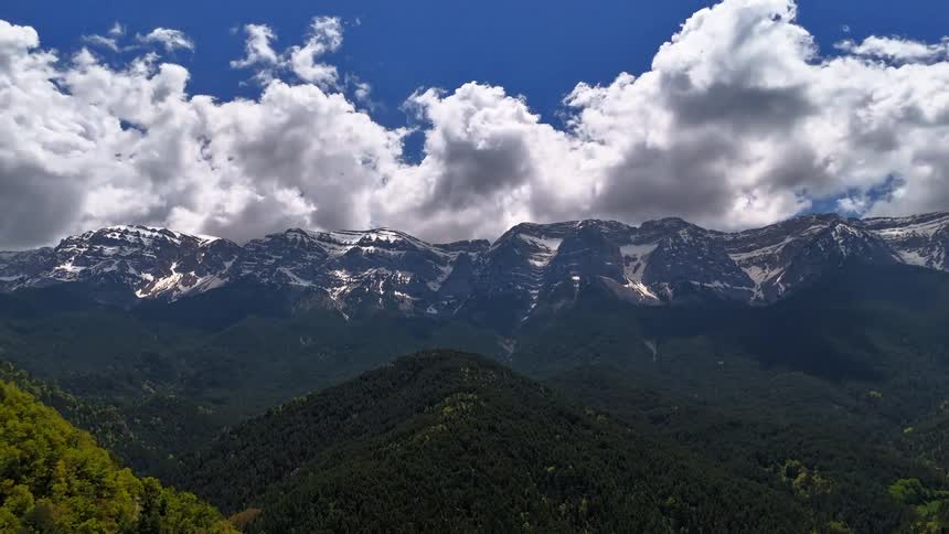 A majestic snow-capped mountain range stands tall under a vibrant blue sky with fast-moving fluffy clouds