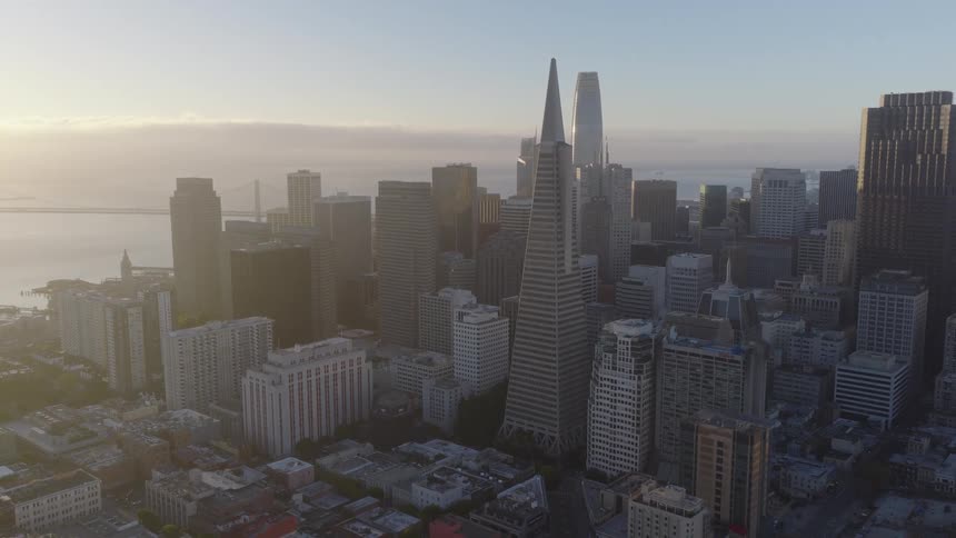 A majestic aerial pullback of the iconic San Francisco skyline with the Transamerica Pyramid and Salesforce Tower glowing in the hazy sunrise light