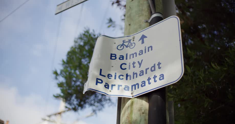 A low-angle close-up of a bent bicycle path sign on a utility pole pointing towards Balmain, City, Leichhardt, and Parramatta in Sydney, Australia