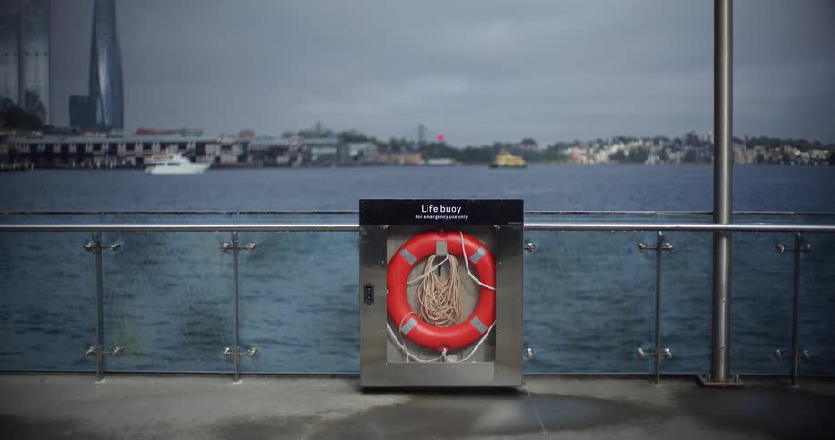 A red life buoy in a case sits on a city pier overlooking the harbor on a cloudy day