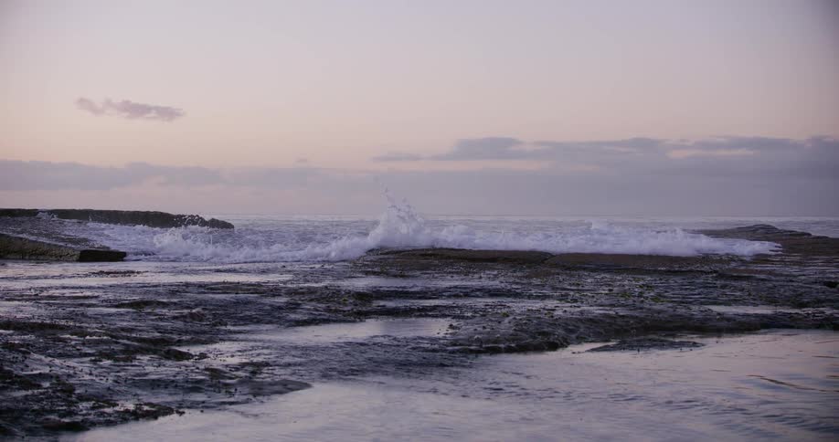 A powerful ocean wave crashes and explodes against a rocky shore in dramatic slow motion during a beautiful, soft dusk