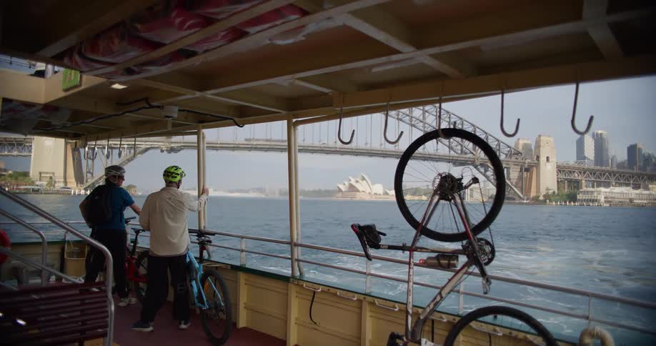 Two cyclists on a ferry admire the scenic view of the Sydney Harbour Bridge and Opera House