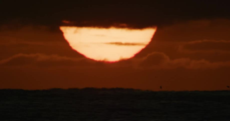 Close-up telephoto view of the fiery sun setting behind dark clouds over a calm ocean as birds fly in silhouette