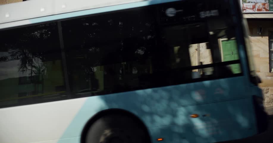 A public transit bus drives in slow motion up a narrow street between historic sandstone buildings