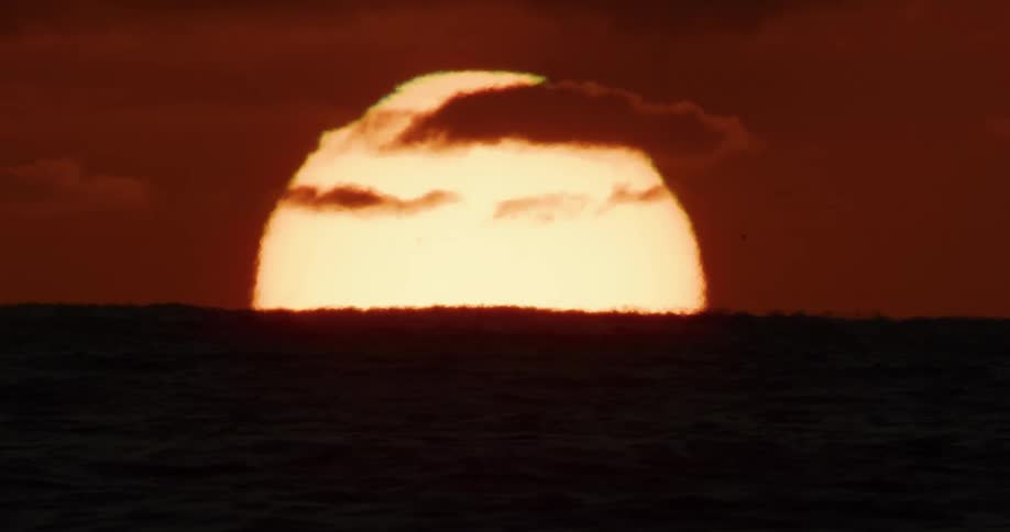 A dramatic close-up of the large sun setting over the dark ocean horizon under a fiery red sky