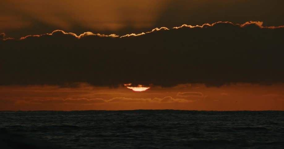 The sun peeks through a sliver between dark clouds over the ocean during a dramatic golden sunset