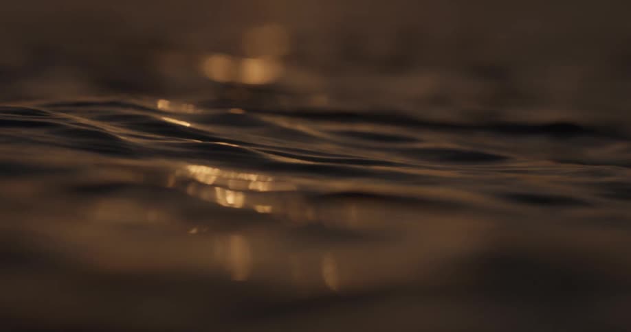 A close-up, slow-motion shot of gentle water waves shimmering with golden sunlight during sunset